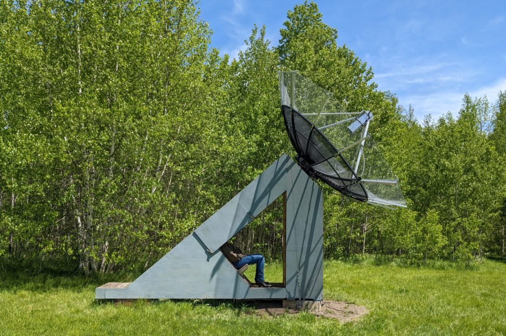 Here GOES Radiotelescope installation at Wave Farm. An 11-foot diameter satellite dish has a seat within a wooden structure beneath it. A seated person looks up at the back of the satellite dish, and are in line with the distant satellite. Seated viewers see a monitor with images being live-transmitted by the geostationary NOAA weather satellite, GOES-16.