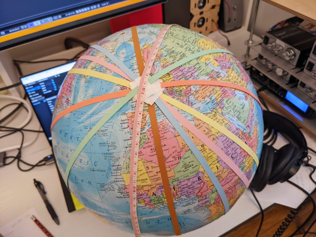 A globe sitting on a desktop with thin paper tapes wrapped around it in different directions, a mockup indicating orbit paths